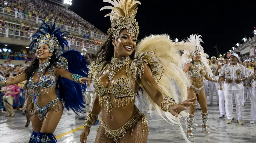 Erwachsene Sambatänzerinnen in glitzernden, federreichen Karnevalskostümen im Vordergrund einer Parade in Rio de Janeiro